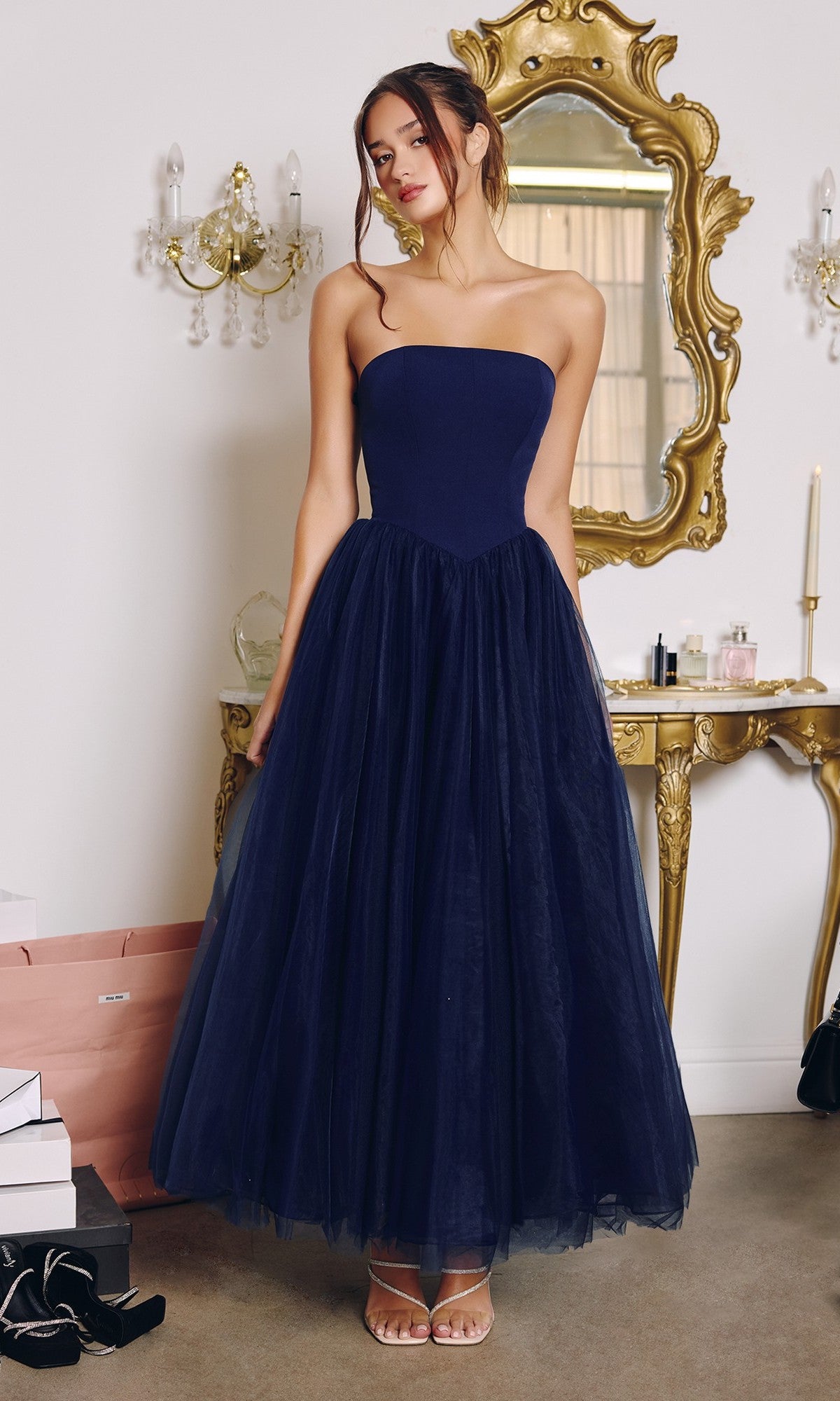 A woman wearing the Classic Strapless Prom Ball Gown: Amoris T712 in navy stands before a gold ornate mirror and vanity table, shopping bags and boxes at her feet.